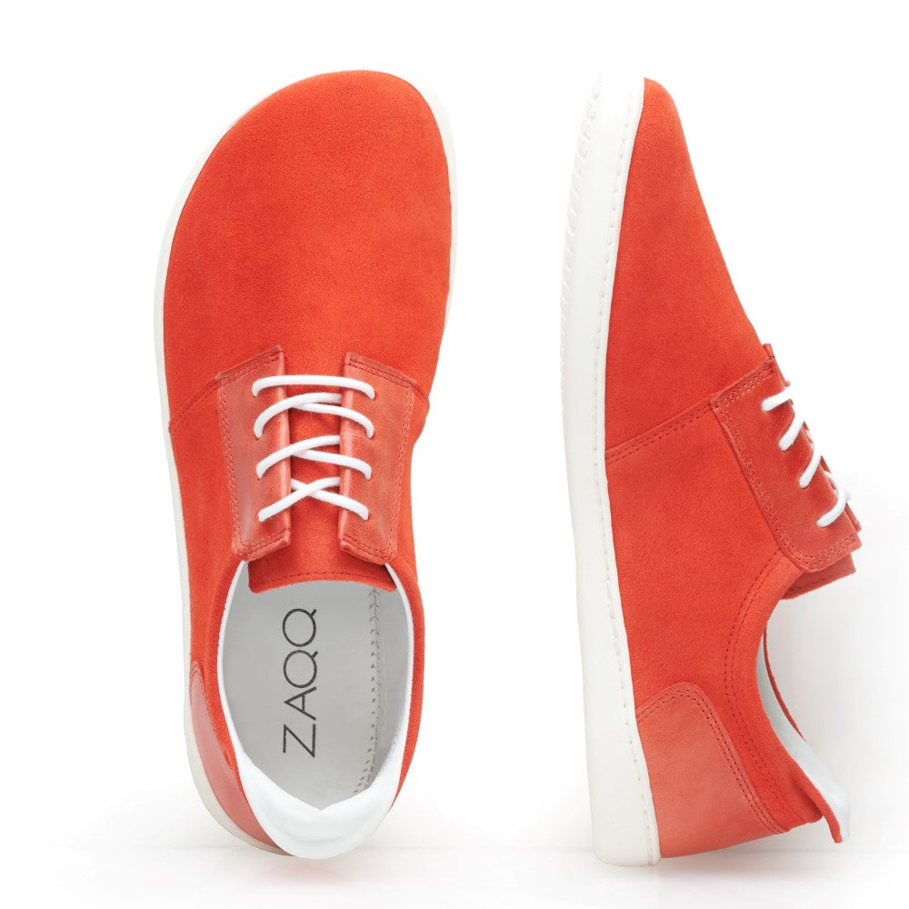 Ein Paar leuchtend orange Wildleder-Sneaker mit weißen Schnürsenkeln und Sohlen im PIQUANT Opal-Design von ZAQQ. Ein Schuh steht aufrecht, während der andere auf der Seite liegt und den Markennamen im Inneren zeigt. Diese modischen Barfußschuhe stehen elegant vor einem schlichten weißen Hintergrund.