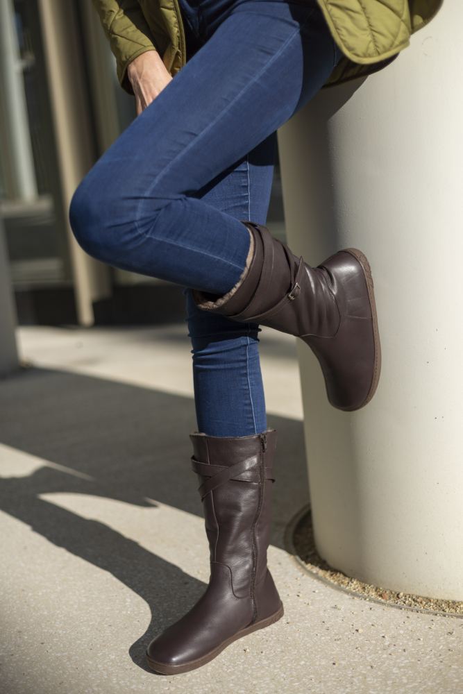 Bekleidet mit ATTRAQT Winter Brown-Stiefeln und Blue Jeans verkörpert die Person Tragekomfort, während sie lässig mit übereinandergeschlagenen Beinen an einer Säule lehnt. Im Freien in einer grünen Jacke ist sie teilweise von der Seite sichtbar, während helles Sonnenlicht Schatten auf den Boden wirft und ihre Wahl des ZAQQ-Schuhwerks hervorhebt.