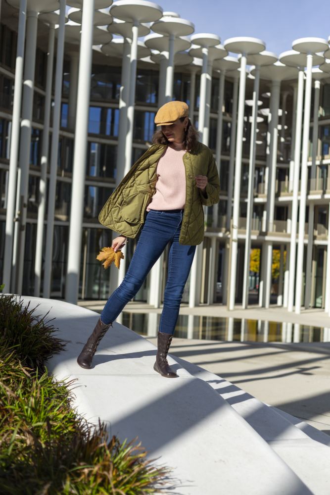 Eine Person in Khakijacke, rosa Pullover und Bluejeans steht draußen auf dem Betonsims eines modernen Gebäudes und trägt die ATTRAQT Stretch Nappa Brown-Stiefel von ZAQQ. Sie hält ein gelbes Ahornblatt in der Hand und trägt eine braune Schiebermütze.