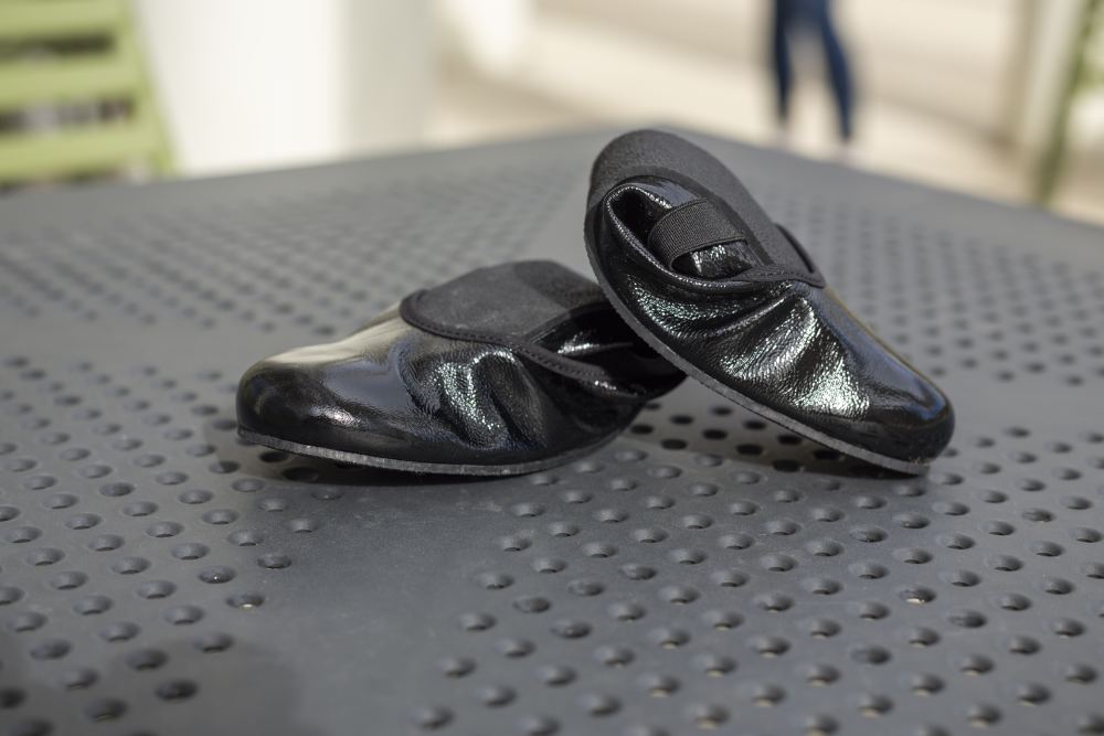 Ein Paar glänzende schwarze TWIST CASEMIRO Flats von ZAQQ ruhen auf einer perforierten Metalloberfläche. Mit elastischen Riemen und einem kleinen Absatz versprechen diese bequemen Schuhe mit ihrer flexiblen Sohle vor einem sanft verschwommenen Hintergrund Komfort.