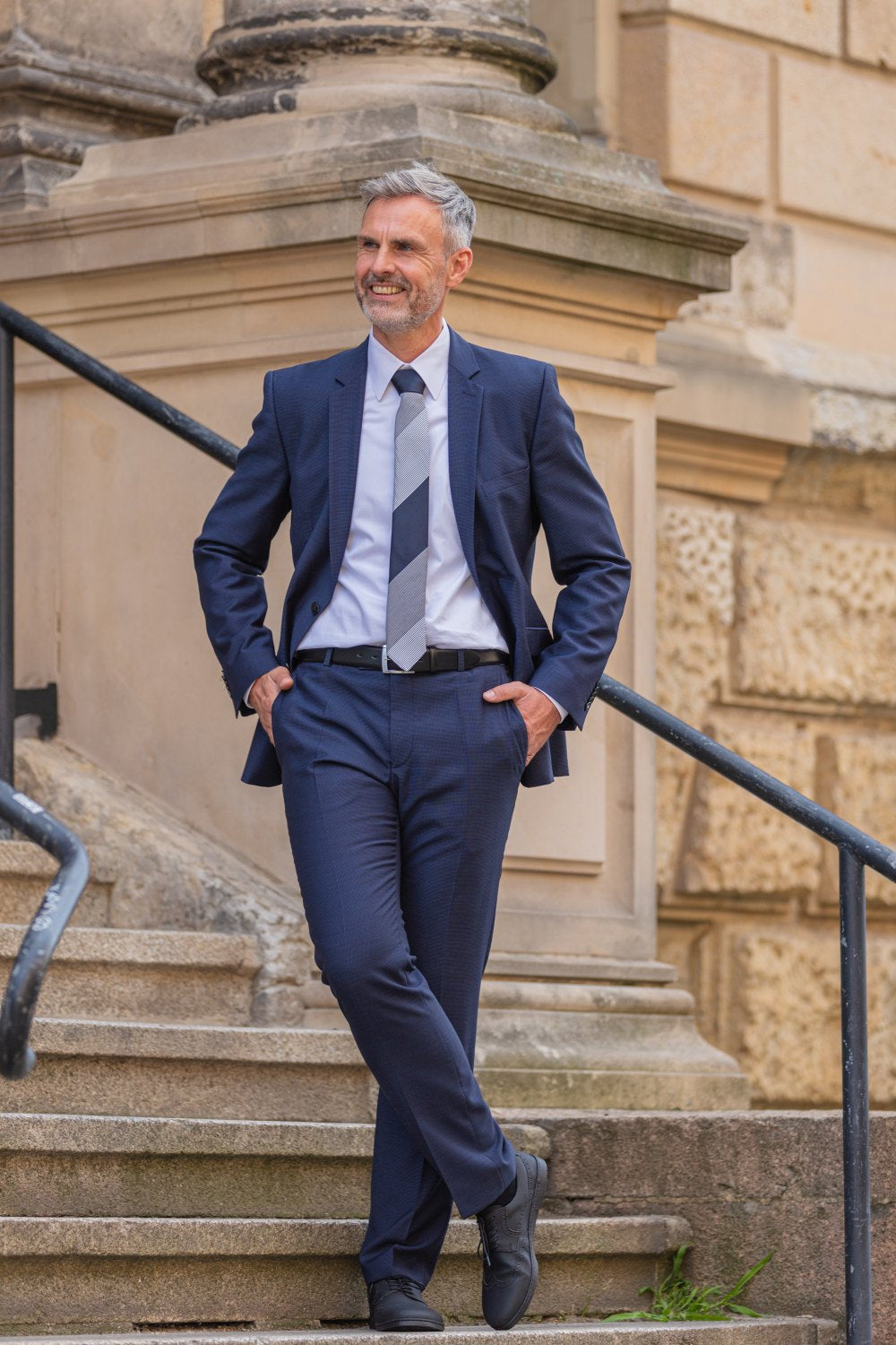 Ein Mann im marineblauen Anzug lehnt lässig an einem Steingebäude und lächelt. Sein graues Haar und sein ordentlich gestutzter Bart tragen zu seinem vornehmen Aussehen bei. Er trägt ZAQQs BRIQ Brogue Vegan Black-Schuhe mit rutschhemmender Sohle und hat seine Hände in den Taschen auf einer Treppe mit verziertem Mauerwerk und einem Metallgeländer.