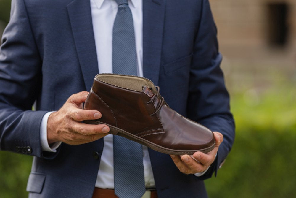 Eine Person im Anzug hält den Schuh DUQE Vegan Antique Brown von ZAQQ, der aus veganen Materialien hergestellt ist, und steht im Freien mit viel Grün im Hintergrund.