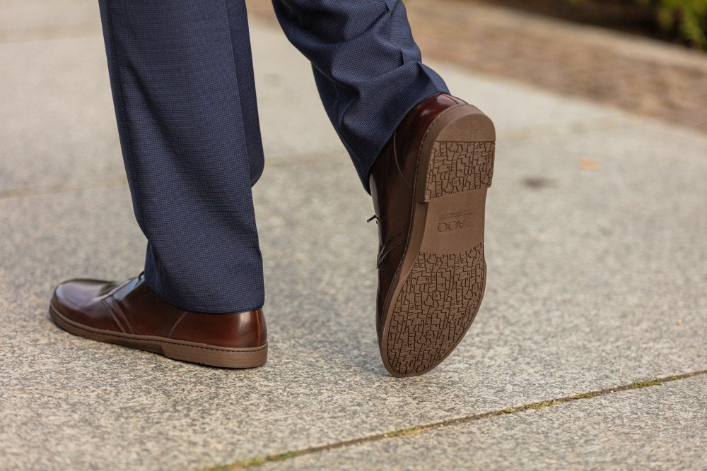 Eine Person in blauer Hose und Schuhen ZAQQ DUQE Vegan Antique Brown läuft auf einem Steinweg. Die Sohle eines Schuhs ist sichtbar und weist ein kompliziertes Muster auf.