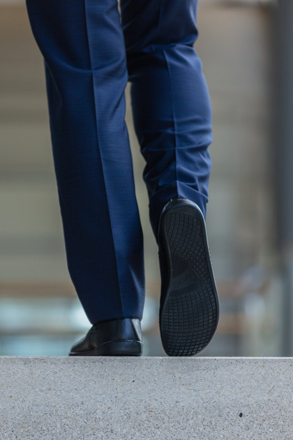 Eine Person, bekleidet mit dunkelblauen Hosen und einem Paar EQUITY Brogue Black von ZAQQ, steigt auf eine Betonfläche. Das Highlight ist die hochwertige Nappaledersohle des linken Schuhs, während der Hintergrund undeutlich bleibt und auf eine urbane Umgebung hindeutet.