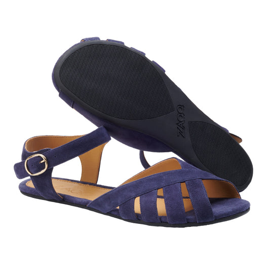 Ein Paar QORSE Blue Sandalen von ZAQQ, aus marineblauem Veloursleder, mit flachen Sohlen, Knöchelriemen und einem Cut-out-Design. Eine Sandale steht aufrecht und zeigt die Draufsicht und die Seitenansicht, während die andere umgedreht ist und ihre strukturierte schwarze Sohle zeigt. Diese Barfußsandalen verbinden nahtlos Stil mit Komfort.