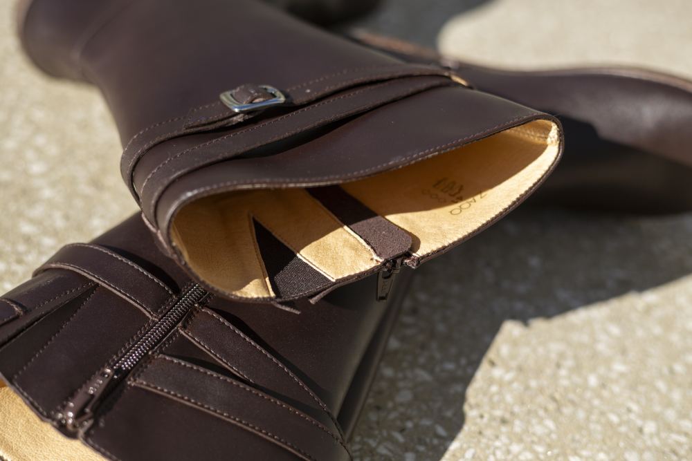 Nahaufnahme eines Paars schwarzer Lederstiefel, die auf dem Boden liegen. Ein Stiefel ist teilweise geöffnet und gibt den Blick auf das beigefarbene Innenfutter frei. Diese ATTRAQT Vegan Brown-Stiefel von ZAQQ haben einen Reißverschluss und einen dekorativen Riemen mit einer Metallschnalle. Das Licht wirft weiche Schatten und hebt die flexible Sohle für zusätzlichen Komfort hervor.