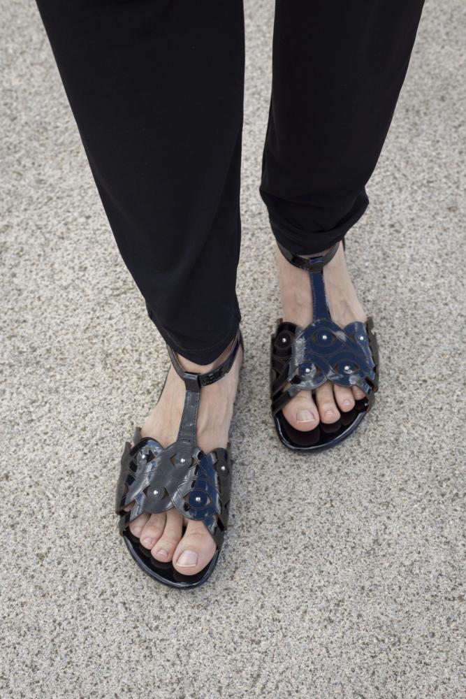 Eine Person, die schwarze Sandalen mit Metallnieten von ZAQQ BOUQET trägt, steht auf einer strukturierten Oberfläche. Das Schuhwerk im Gladiator-Stil aus glattem Lackleder passt perfekt zu der schwarzen Hose.