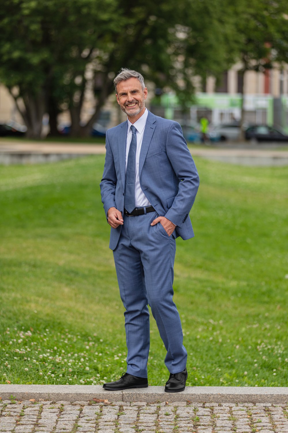 Ein Mann im blauen Anzug steht selbstbewusst auf einem Steinweg in einem Park. Seine BRIQ Brogue Black-Schuhe von ZAQQ verleihen ihm eine elegante Note. Mit einer Hand in der Tasche und einem Lächeln im Gesicht wird die Szene von grünem Gras, Bäumen und verschwommenen Gebäuden eingerahmt.