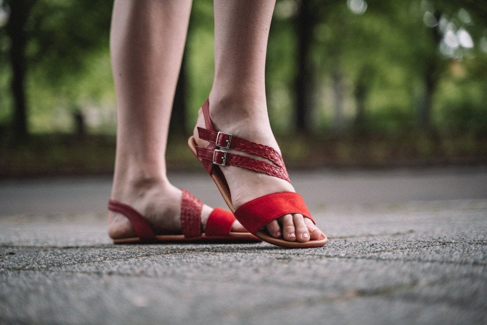 Nahaufnahme einer Person, die die roten BROOQE Sandalen von ZAQQ mit zwei Schnallen aus Veloursleder trägt und auf einer gepflasterten Fläche steht. Der Hintergrund zeigt eine verschwommene grüne Außenkulisse, die den außergewöhnlichen Tragekomfort der Sandalen hervorhebt.