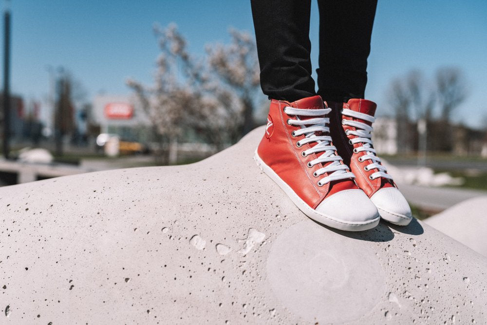 Eine Person, die rote High-Top-Sneakers von ZAQQ mit weißen Schnürsenkeln trägt, steht selbstbewusst auf einem großen weißen Felsen. Die verschwommene Außenszene mit Bäumen und klarem blauen Himmel bietet eine erfrischende Kulisse für diese lebendigen Sneaker aus geschmeidigem Nappaleder.