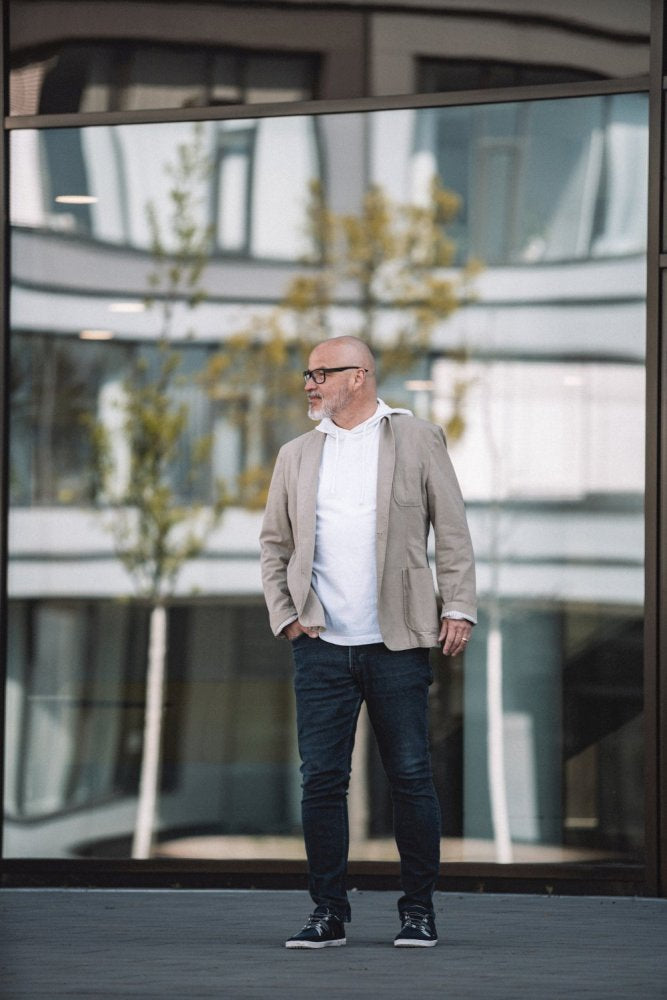 Ein Mann mit Bart und Brille steht selbstbewusst vor einem Glasgebäude. Er trägt eine beige Jacke, ein weißes Hemd, Jeans und DRAQE Azul von ZAQQ, bekannt für ihren außergewöhnlichen Tragekomfort. In der Spiegelung des Gebäudes sind Bäume und architektonische Details zu sehen.