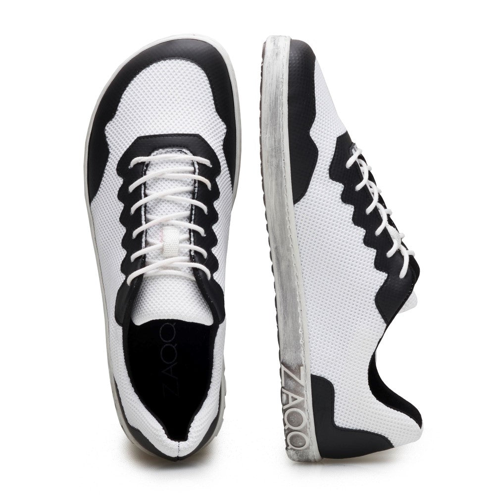 Wir präsentieren die DUPLEQ Black White Vegan-Sneaker von ZAQQ. Diese stylischen veganen Barfußschuhe haben ein atmungsaktives, luftiges Obermaterial aus weißem Mesh mit schwarzen Akzenten. Das Design umfasst weiße Schnürsenkel und eine graue Sohle für zusätzlichen Komfort. Ein Schuh ist von oben zu sehen, was seinen Schnürstil hervorhebt, während der andere das markante ZAQQ-Logo an der Seite hervorhebt.