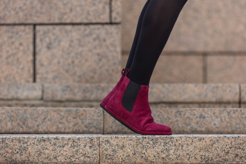 Eine Person trägt ZAQQs EQUITY Berry Chelsea Boots aus rotem Wildleder mit schwarzen elastischen Seiteneinsätzen und geht Steintreppen hinauf. Diese stilvollen Stiefel, die an die Mischung aus Glattleder und Wildleder erinnern, ergänzen den Hintergrund einer Steinmauer mit sichtbaren Nähten perfekt.