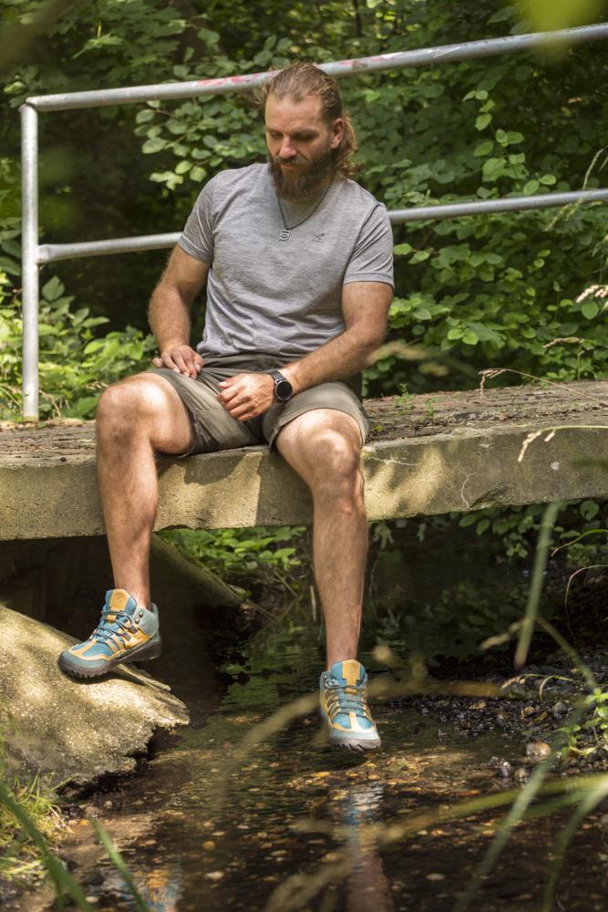 Ein Mann mit Bart, grauem Hemd und Shorts sitzt auf einer kleinen Betonbrücke über einem Bach. Seine wasserdichten Schuhe ZAQQ ESQAPE Blue Orange, die so bequem wie robust sind, baumeln über dem Wasser. In Gedanken versunken inmitten des üppigen Grüns glitzert seine treue Uhr im Sonnenlicht.