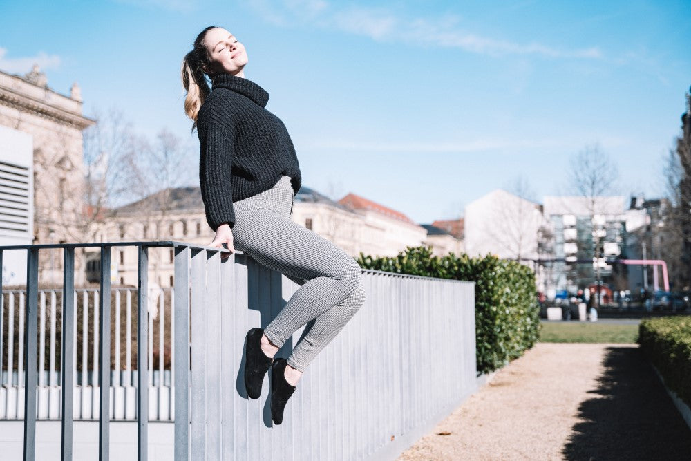 Eine Frau in schwarzem Pullover und karierter Hose lehnt sich auf einem Geländer zurück und genießt den sonnigen Tag in ihren bequemen ZAQQ IQON Velours Black Schuhen mit ihrem weichen Veloursfinish. Sie lächelt mit geschlossenen Augen, während Gebäude und Grünflächen eine ruhige Kulisse unter dem weiten blauen Himmel bilden.