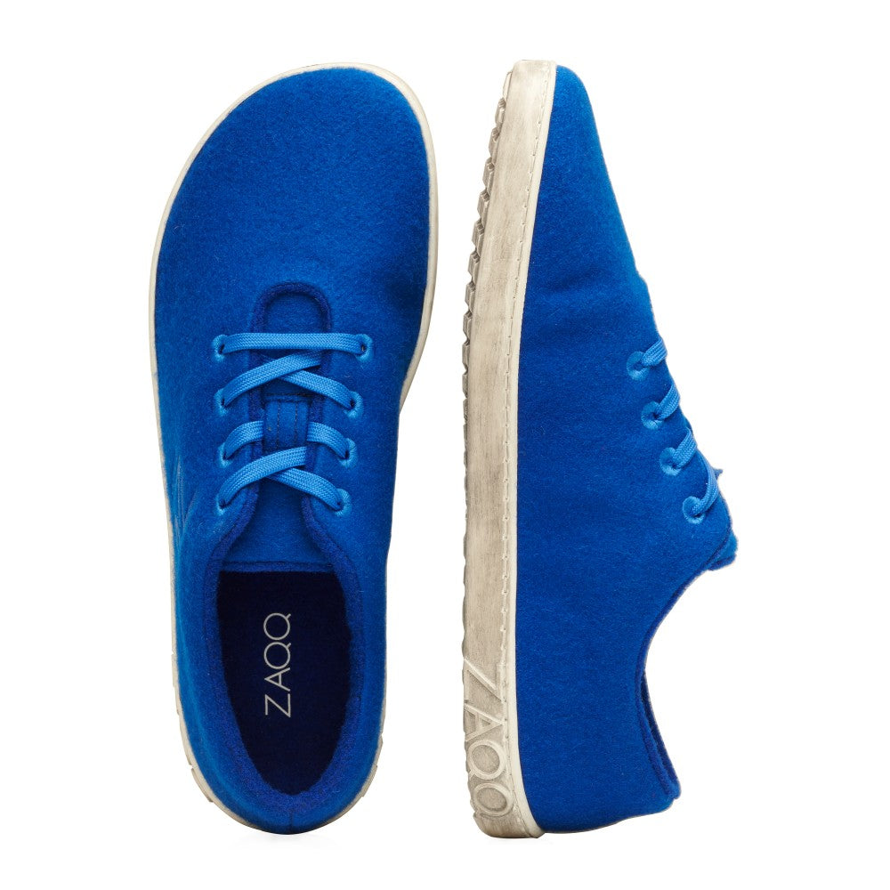 Ein attraktives Paar LIQE Azur-Sneaker von ZAQQ aus Merinowolle zeigt eine leuchtend blaue Farbe, wobei ein Schuh von der Seite und der andere von oben zu sehen ist. Diese Schuhe haben weiße Sohlen und blaue Schnürsenkel, während die Innensohle des oberen Schuhs stolz ihr Branding als einzigartiger Barfußschuh zeigt.