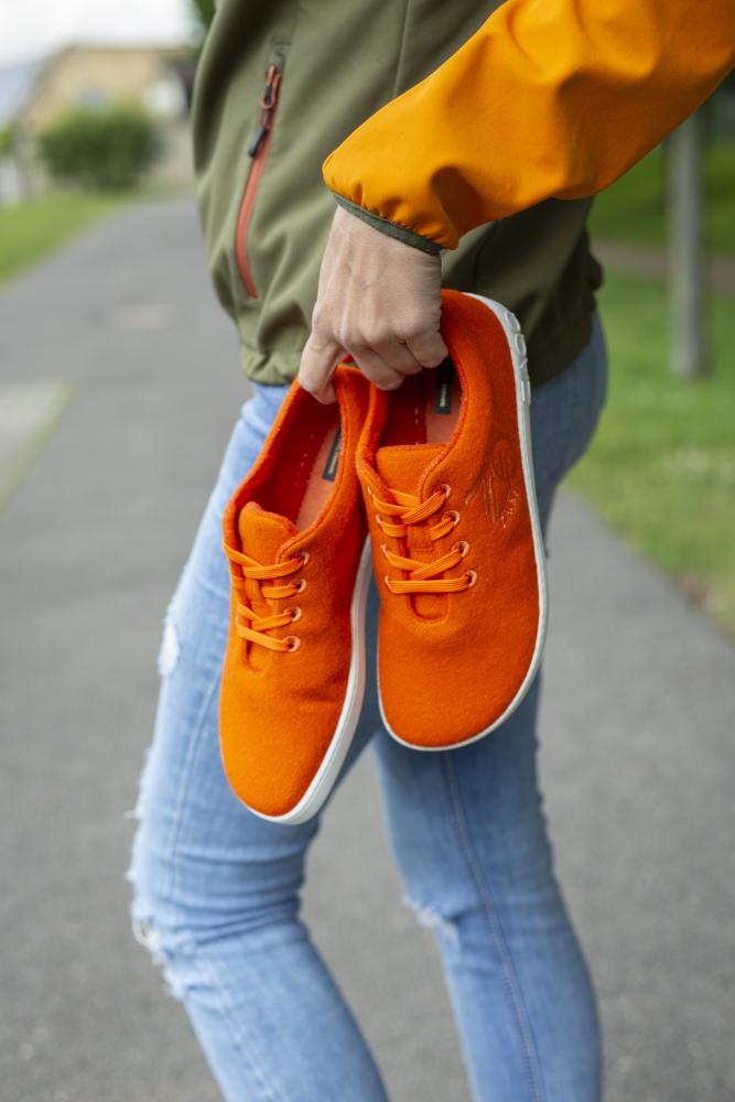 Eine Person in einer grün-orangen Jacke und blauen Jeans hält die leuchtend orangefarbenen LIQE-Sneaker von ZAQQ, die ein Barfußgefühl vermitteln sollen, während sie auf einem gepflasterten Weg steht.