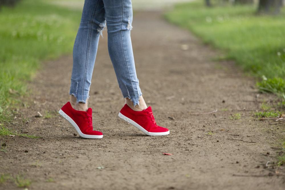 Eine Person, die ZAQQs LIQE Race-Sneaker mit Barfußsohle und zerrissene Jeans trägt, schlendert einen Feldweg entlang, der beiderseits von Gras bedeckt ist.