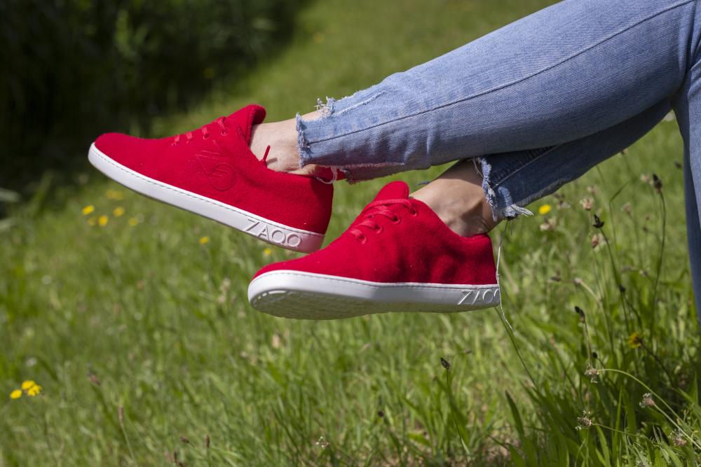 Eine Person in LIQE Race Sneakers von ZAQQ und zerrissenen Bluejeans sitzt mit überkreuzten Beinen über dem üppigen Gras. Im Hintergrund ist eine Grünfläche mit kleinen gelben Blumen zu sehen, die der ruhigen Szene einen frischen Farbtupfer verleihen.