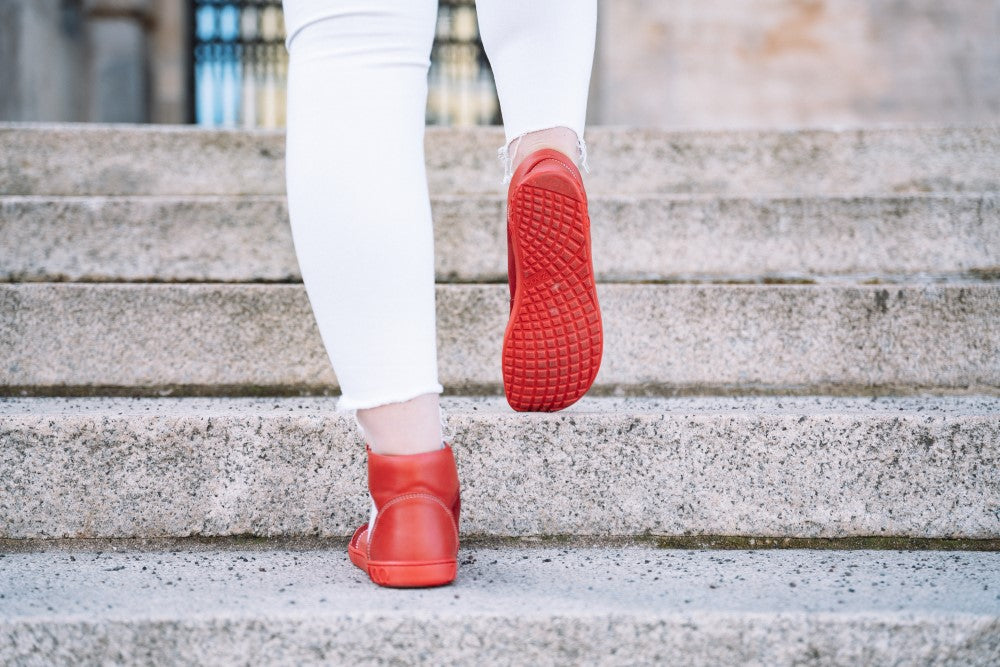 Eine Person in weißer Hose und einem Paar LUQQ Red Sneakers von ZAQQ mit flexibler Kautschuksohle geht eine Steintreppe hinauf. Der Fokus liegt auf den roten Sohlen und der Rückansicht der Schuhe vor den grauen Stufen.