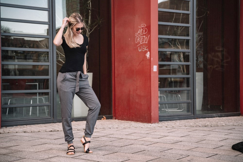 Eine Person mit Sonnenbrille, einem NIQ Black T-Shirt von ZAQQ und grauen Hosen, ergänzt durch moderne Sandalen für maximalen Komfort, steht auf einem gepflasterten Gehweg vor einem modernen Gebäude mit großen Fenstern. Eine rote Wand mit teilweise sichtbarem Graffiti auf der rechten Seite verleiht der Szene einen Kontrast.