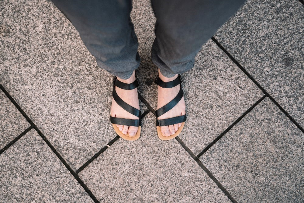 Blick auf die Füße einer Person in den NIQ Black-Sandalen von ZAQQ aus hochwertigem Nappaleder, die auf einem mit Steinfliesen ausgelegten Bürgersteig steht. Das elegante schwarze Riemchendesign bietet maximalen Komfort und ergänzt die dunkle Hose perfekt.