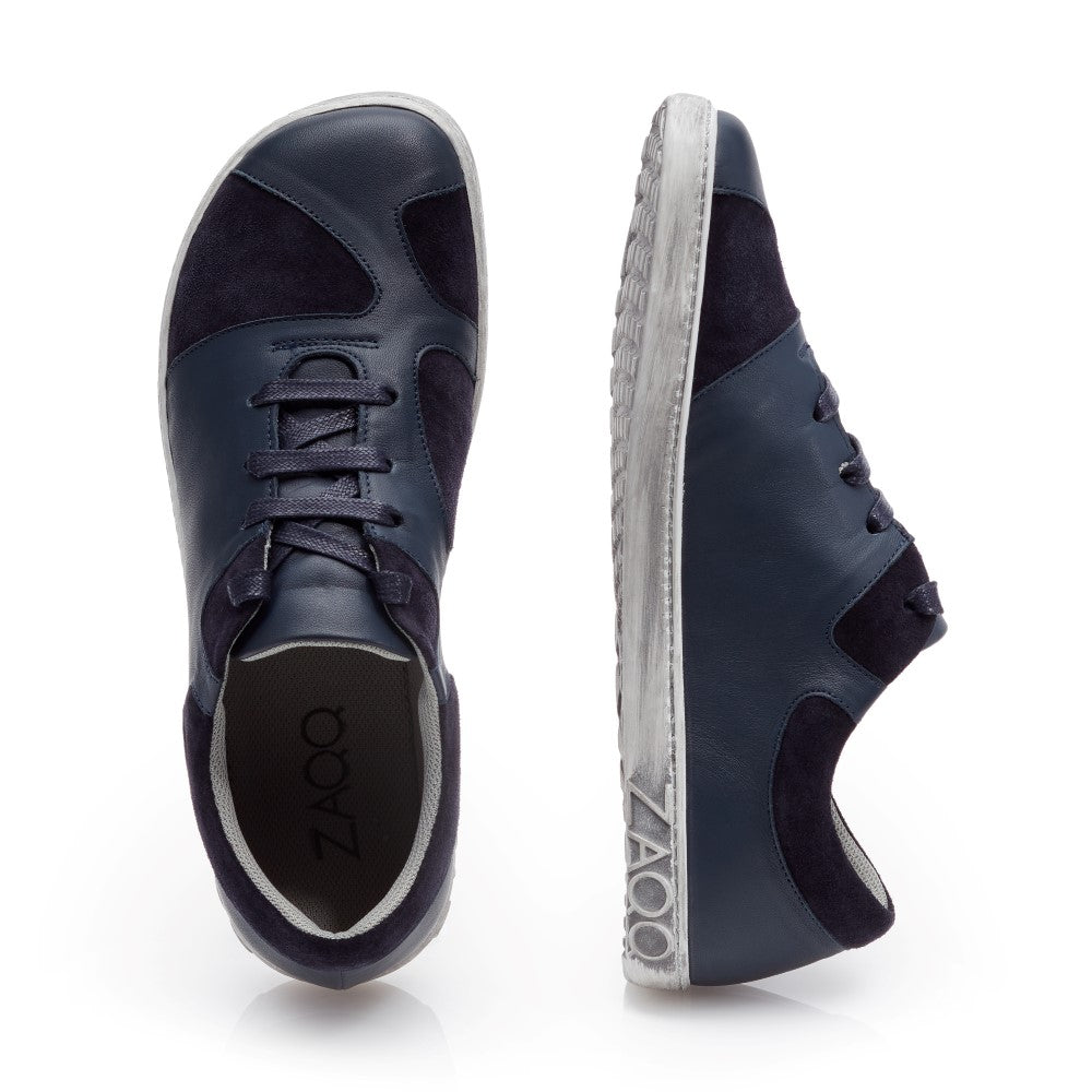 Hier werden die QAANAAQ Blue Sneaker von ZAQQ präsentiert. Ein Schuh aus elegantem Veloursleder steht aufrecht in der Seitenansicht und hat eine weiße Sohle mit dem ZAQQ-Logo. Der andere Schuh ist von oben zu sehen, was sein Schnürdesign und sein schlichtes Finish hervorhebt.