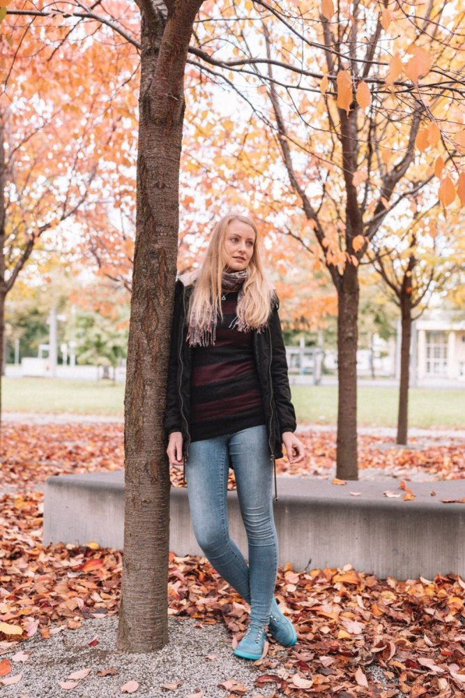 Eine Frau mit langen blonden Haaren steht im Herbst neben einem Baum in einem Park. Sie trägt eine schwarze Jacke, ein gestreiftes Hemd und Jeans, dazu die türkisfarbenen QARLY-Sneakers von ZAQQ, die sich von den bunten, gefallenen Blättern abheben, die über den Boden verstreut sind.