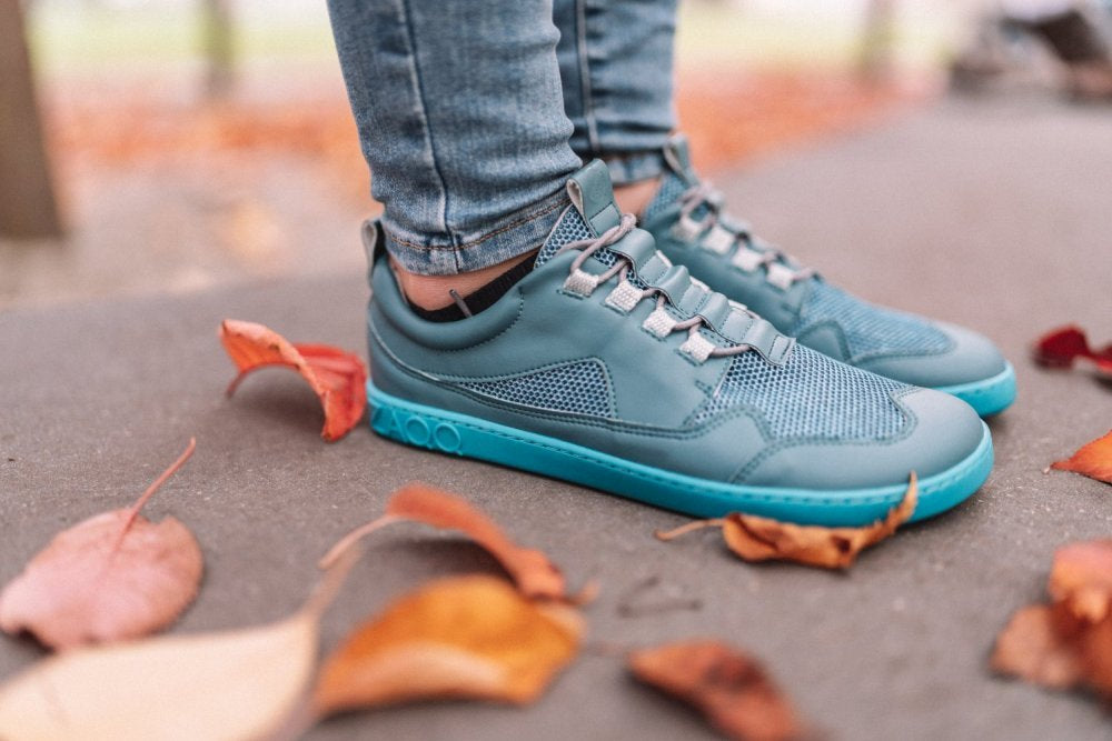 Eine Person steht auf einem mit Herbstlaub geschmückten Weg und trägt den QARLY Turquoise von ZAQQ mit türkiser Sohle und Mesh-Gewebe. Die hochgekrempelte Jeans verleiht dem Schuh einen lässigen Touch, der Hintergrund bleibt leicht unscharf.