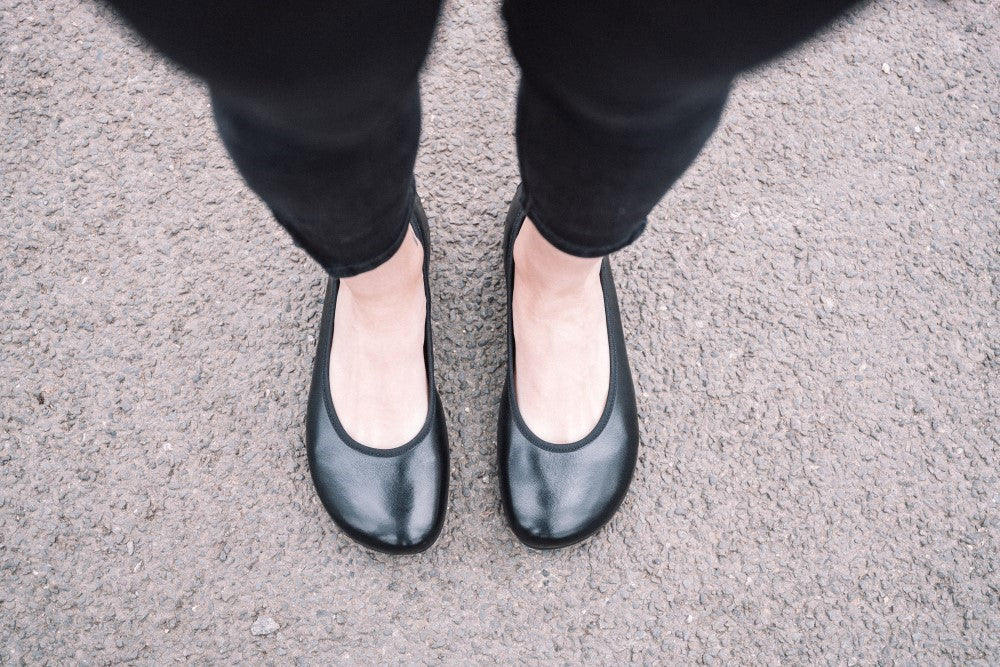 Eine Person trägt schwarze Ballerinas im Ballerina-Design QARMA von ZAQQ und eine schwarze Hose und steht auf einer strukturierten grauen Oberfläche. Das Bild, das die Eleganz ihrer Haltung aus einer Draufsicht einfängt, hebt das einzigartige, die Passform verbessernde Fußbett der Schuhe hervor.