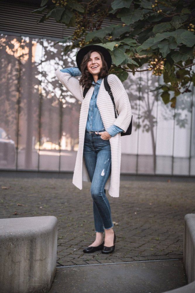 Eine Frau mit breitkrempigem Hut, langer beiger Strickjacke, Jeans-Outfit und eleganten dunkelbraunen QARMA-Schuhen von ZAQQ steht lächelnd im Freien, umgeben von grünen Blättern. Sie steht auf einer gepflasterten Fläche mit moderner Architektur im Hintergrund.