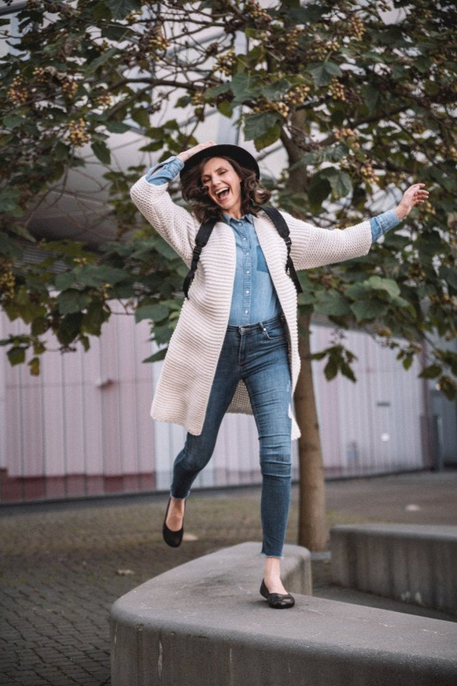 Eine fröhliche Person mit Hut und langer Strickjacke geht voller Energie an einer geschwungenen niedrigen Mauer entlang, umgeben von Bäumen mit grünen Blättern. Sie trägt ein Jeans-Outfit und die dunkelbraunen QARMA-Schuhe von ZAQQ, die an Barfuß-Ballerinas erinnern. Sie trägt einen Rucksack. Die Außenfassade des Gebäudes bietet eine faszinierende Kulisse.