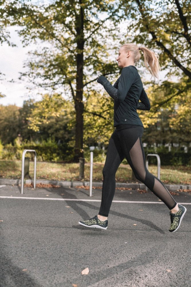 Eine Person mit Pferdeschwanz joggt auf einem gepflasterten Weg in einem Park, umgeben von Bäumen mit grünen Blättern. In einem schwarzen Outfit und QASEY Green Sneakers von ZAQQ aus veganen Materialien genießen sie die helle Szene, die durch das natürliche Licht noch verstärkt wird.