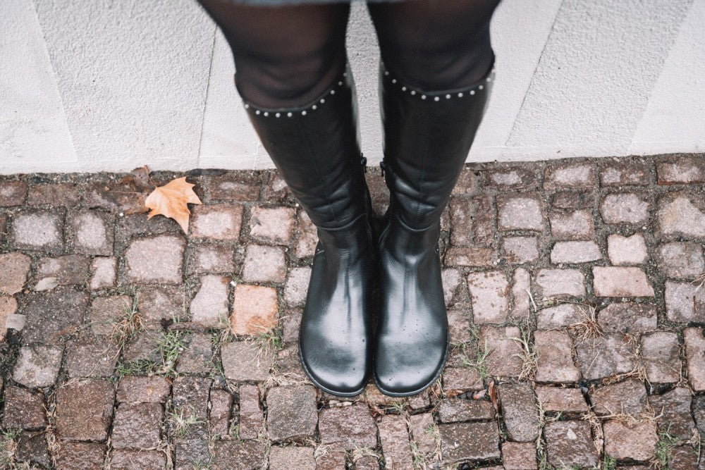 Eine Person, die QATCH SHINE Nappa Black von ZAQQ trägt, steht auf einem Kopfsteinpflasterweg, neben dem sich ein einzelnes braunes Blatt befindet. Die Stiefel haben kleine Stollen am oberen Ende und eine Kautschuksohle für zusätzliche Haltbarkeit, vor einem hellen Wandhintergrund.