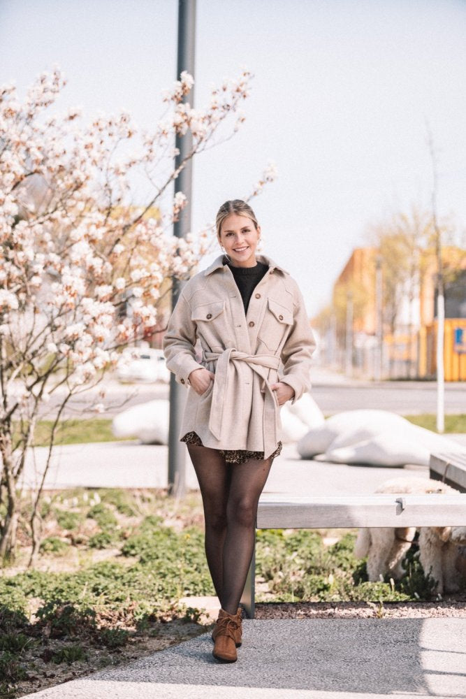 Eine lächelnde Frau in einer beigen Jacke mit Gürtel steht im Freien. Sie trägt ein schwarzes Oberteil und einen gemusterten Rock. Ihre QEBEQ Cognac-Stiefel von ZAQQ ergänzen ihren Look perfekt an diesem sonnigen Tag mit blühenden Bäumen und einem Weg, der sich hinter ihr erstreckt.