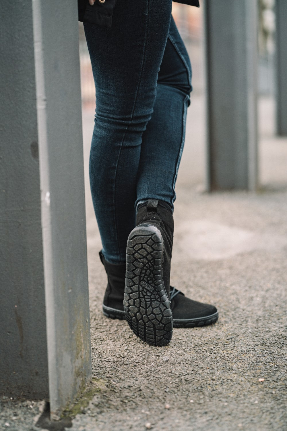 Eine Person in schwarzen QICE-Stiefeln und dunklen Jeans läuft auf einem rauen Bürgersteig. Das Mikrofaser-Detail der Sohle eines Stiefels ist zu sehen, während sie an einem grauen Metallpfosten vorbeigeht, der die Marke ZAQQ repräsentiert.