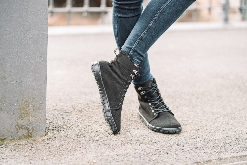 Eine Person, die die veganen High-Top-Sneakers ZAQQ QICE Black mit Schnürsenkeln trägt, steht im Freien und lehnt an einem grauen Pfosten. Der Boden besteht aus Beton und die Person kombiniert die Sneaker mit dunkelblauen Jeans.