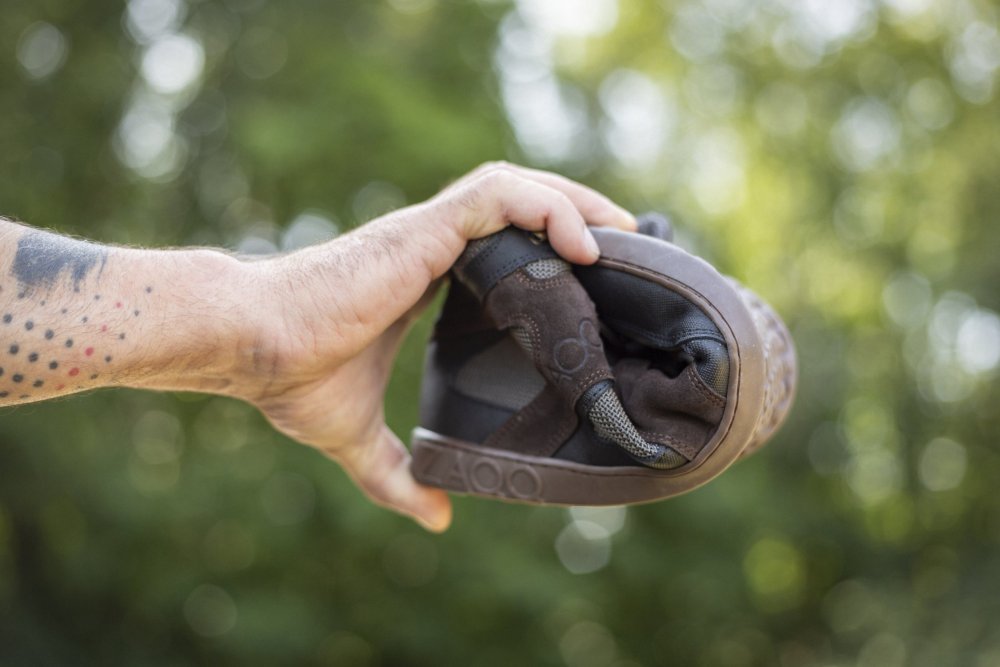 Eine Hand, die den QINSLEY Black von ZAQQ zeigt, anmutig in der Mitte gebogen, um seine Flexibilität zu demonstrieren. Der Schuh besteht aus atmungsaktivem Mesh-Gewebe. Üppiges Grün dient als sanft verschwommener Hintergrund, während auf dem Unterarm ein Tattoo mit kleinen Punkten zu sehen ist, das einen Hauch von persönlichem Stil vermittelt.