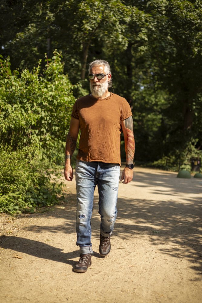 Ein Mann mit Bart und Sonnenbrille schlendert einen sonnenbeschienenen Feldweg im Park entlang. Er trägt ein schwarzes T-Shirt von ZAQQ QINSLEY, zerrissene Jeans und Stiefel. Die Gegend ist mit üppigen grünen Bäumen und Blättern bewachsen, was eine einladende natürliche Atmosphäre schafft.