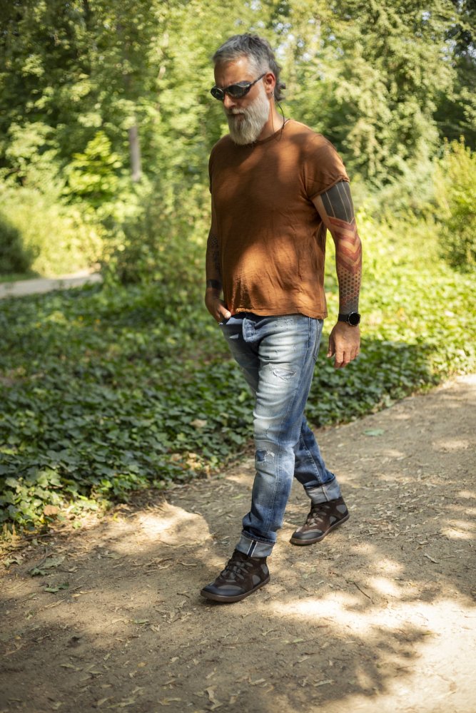 Ein Mann mit Bart und Sonnenbrille geht einen Feldweg in einem Park entlang. Er trägt schwarze QINSLEY-Jeans und Stiefel von ZAQQ und hört mit Kopfhörern Musik. Sein braunes T-Shirt ergänzt die üppige Kulisse aus grünen Bäumen und Büschen und schafft eine ruhige Szene.