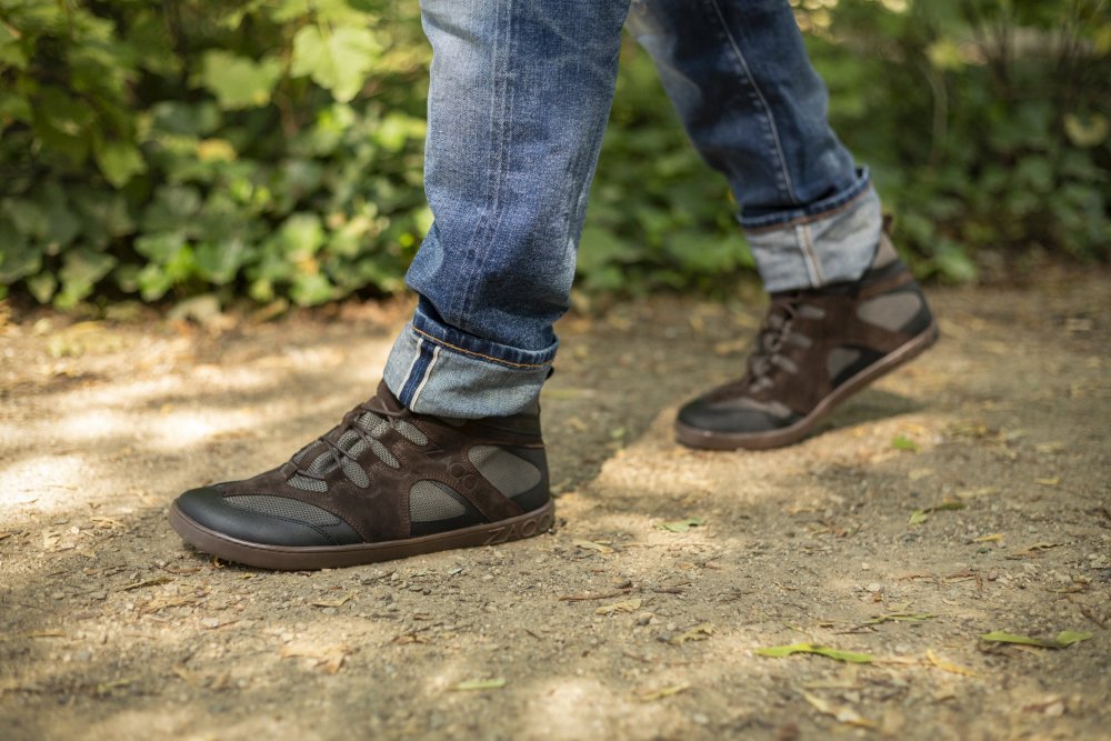 Eine Person in hochgekrempelten Jeans und den Wanderschuhen QINSLEY Black von ZAQQ mit robuster Outdoorsohle läuft auf einem Schotterweg inmitten von Grün. Die Schuhe aus atmungsaktivem Mesh-Gewebe wirken robust und ideal für Outdoor-Abenteuer.