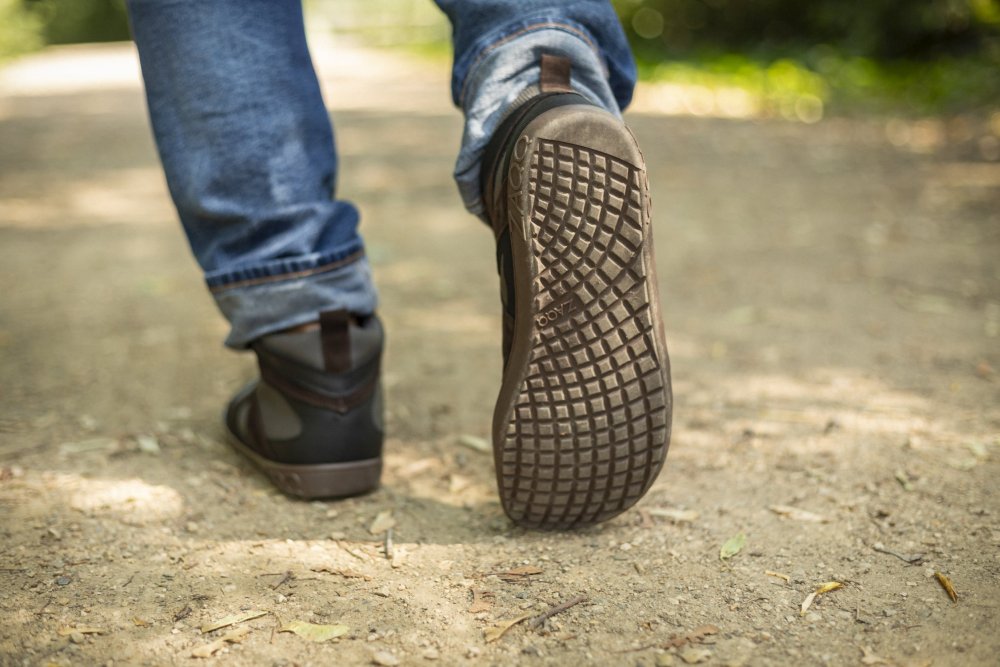 Nahaufnahme des Fußes einer Person, die den schwarzen Barfußschuh ZAQQ QINSLEY mit Outdoorsohle trägt und auf einem Feldweg läuft, auf dem das komplizierte Sohlenmuster sichtbar ist. Die Person trägt blaue Jeans und der Hintergrund zeigt weichgezeichnetes Grün.