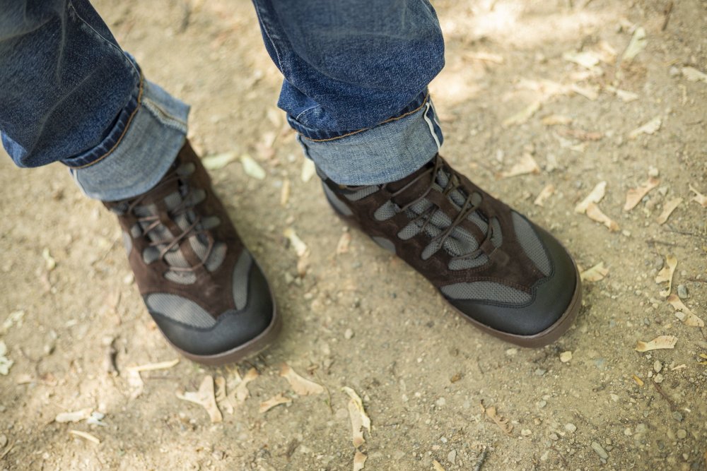 Eine Person in den schwarzen QINSLEY Wanderschuhen von ZAQQ aus atmungsaktivem Meshgewebe und Blue Jeans steht auf einem mit kleinen Blättern übersäten Feldweg. Die Jeans ist über den Schuhen leicht hochgekrempelt und gibt den Blick auf die robuste ZAQQ Outdoorsohle frei.