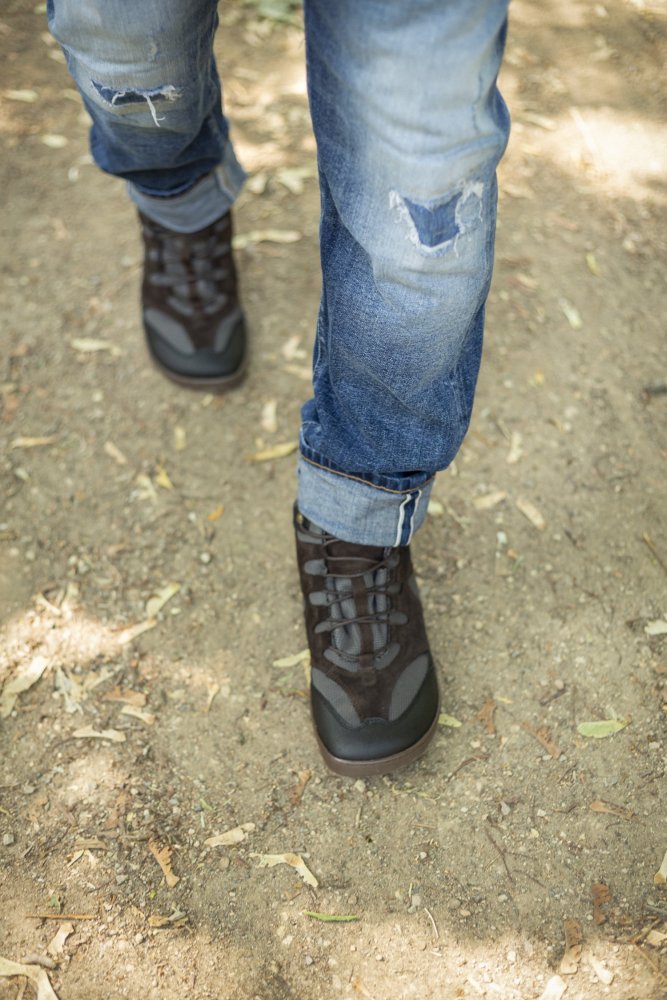 Eine Person schlendert einen Feldweg entlang und trägt schwarze QINSLEY-Sneaker von ZAQQ aus atmungsaktivem Netzgewebe und hochgekrempelte zerrissene Jeans. Sonnenlicht fällt durch die Bäume und wirft Schatten auf den Boden, während gefallene Blätter verstreut herumliegen.