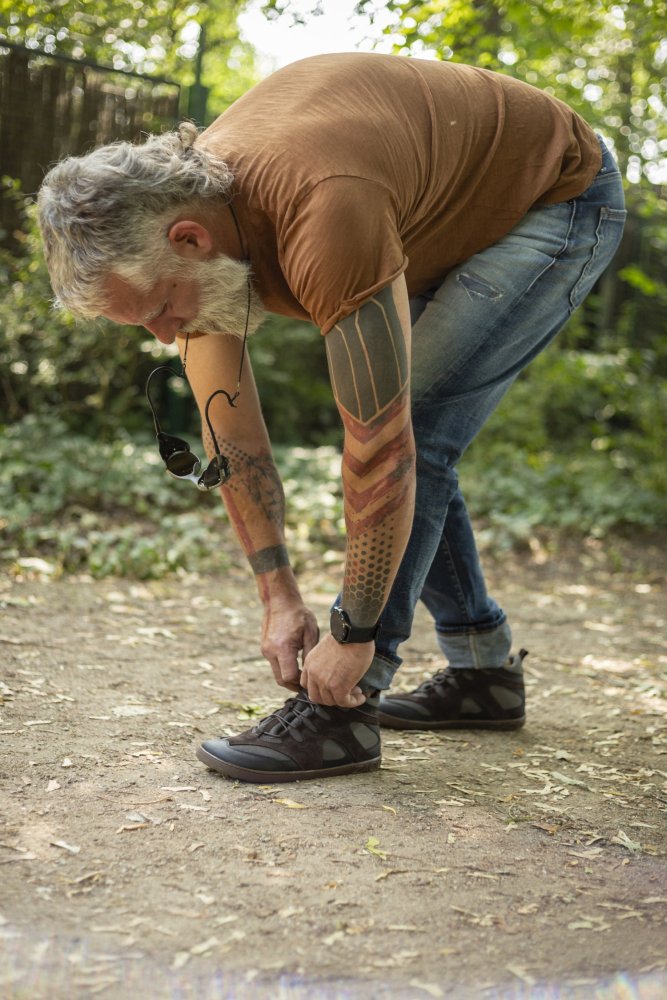 Ein bärtiger Mann mit Tattoos bückt sich, um auf einem Feldweg im Wald seine Schnürsenkel zu binden, und schlüpft in seine schwarzen QINSLEY-Schuhe von ZAQQ mit ihrer robusten Outdoor-Sohle. Er trägt ein braunes T-Shirt, Jeans und eine Sonnenbrille um den Hals, umgeben von üppigem Grün.