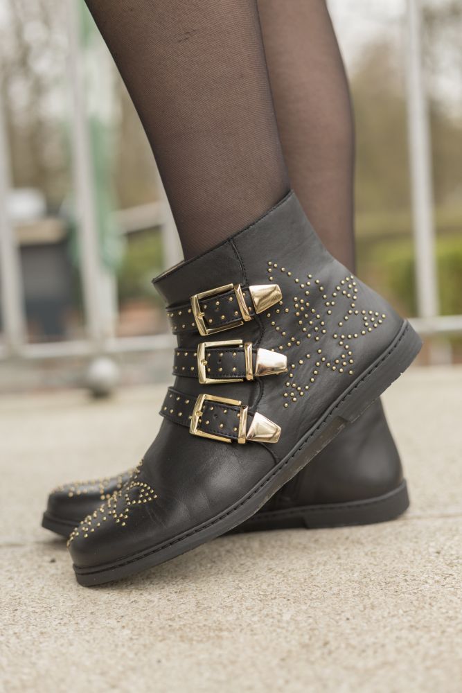 Nahaufnahme einer Person, die die ZAQQ QLANCE Black Gold Stiefeletten trägt, verziert mit goldenen Nieten und drei Schnallenriemen. Diese Stiefel weisen ein kompliziertes Blumenmuster auf, während die Trägerin selbstbewusst vor einem verschwommenen Außenhintergrund steht.