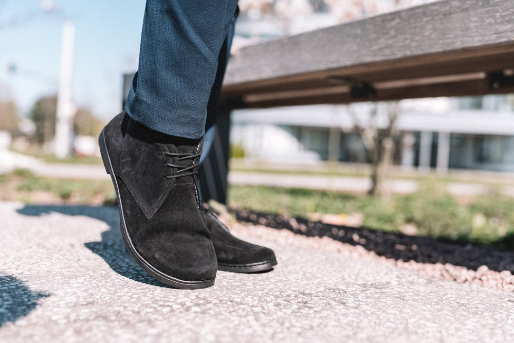 Nahaufnahme einer Person, die schwarze ZAQQ QLASSY-Schuhe und marineblaue Hosen trägt und auf einem Kiesweg läuft. Im Hintergrund sind eine Holzbank und verschwommene Gebäude zu sehen.