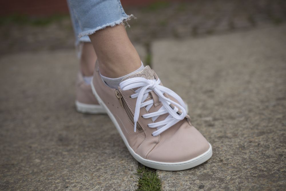 Eine Person trägt den Sneaker QOMFY Wide Rosa von ZAQQ, in hellrosa mit weißen Schnürsenkeln und seitlichem Reißverschluss, und steht auf einem Steinpflaster. Diese Sneaker aus hochwertigem Nappaleder ergänzen die hellblaue Jeans perfekt.