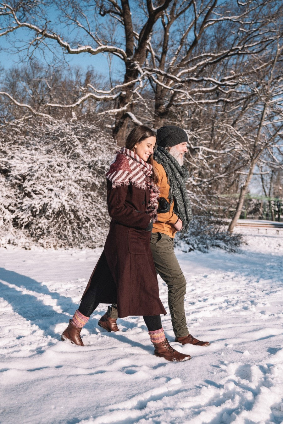 Ein Mann und eine Frau gehen gemeinsam durch einen schneebedeckten Park, eingehüllt in warme Mäntel und Schals, beide tragen QUINTIC Winter Vegan Nut Stiefel von ZAQQ. Die kahlen Bäume und der klare blaue Himmel bieten eine atemberaubende Kulisse für ihren Winterausflug.