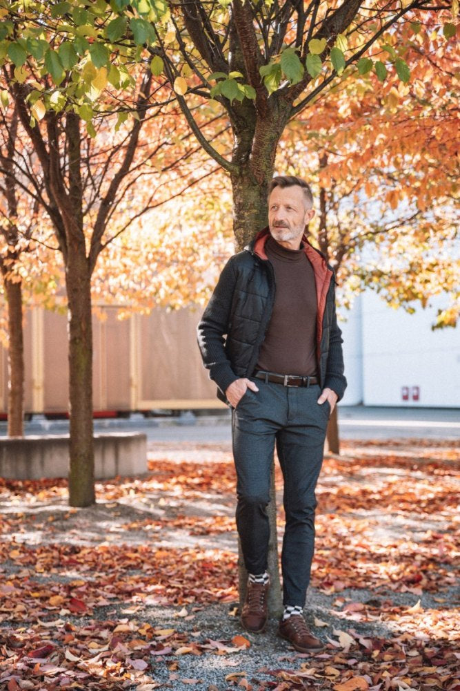 Ein Mann steht entspannt unter einem Baum in einem mit Herbstlaub bedeckten Park und genießt den Komfort seiner braunen ZAQQ RIQO-Schuhe. Er trägt eine dunkle Jacke, ein braunes Hemd und graue Hosen mit braunen Schuhen und genießt das atmungsaktive Gefühl, während orangefarbenes und rotes Laub eine warme, saisonale Kulisse bietet.